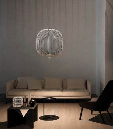 Ein minimalistisches Wohnzimmer mit der Spokes Pendelleuchte von Foscarini als elegantem Mittelpunkt. Die filigranen, metallischen Streben der Leuchte werfen sanfte Lichtspiele an Wand und Decke und verleihen dem Raum eine leichte, skulpturale Wirkung. Helles Sofa, dunkle Akzente und natürliche Materialien schaffen eine ruhige, moderne Atmosphäre.