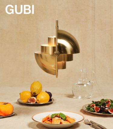 Eine elegante Multi-Lite Pendelleuchte von GUBI in gebürstetem Messing schwebt über einem gedeckten Tisch. Darunter stehen Teller mit Pasta, frischen Feigen und Zitronen sowie eine Glaskaraffe – eine stilvolle Szene, die Design und Genuss vereint.