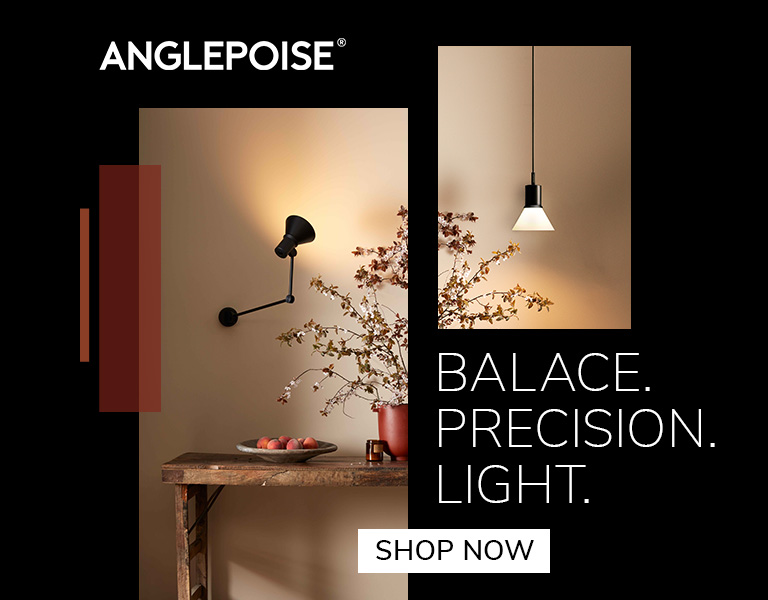 A black Anglepoise wall lamp illuminating a rustic wooden table with a bowl of fruit and a vase of branches. Another minimalist pendant light hangs on a beige wall. Text reads: &lsquo;Anglepoise. Balance. Precision. Light.&rsquo; A button says &lsquo;Shop Now.&rsquo;