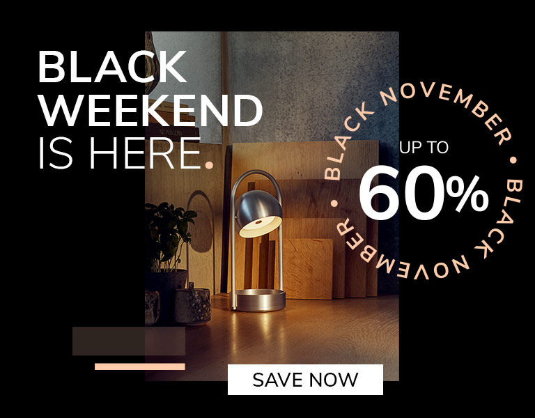Modern silver table lamp glowing on a wooden desk with cutting boards and plants in the background. Promotional text reads: &lsquo;Black Weekend is here. Up to 60%. Black November.&rsquo; A button says &lsquo;Save Now.&rsquo;